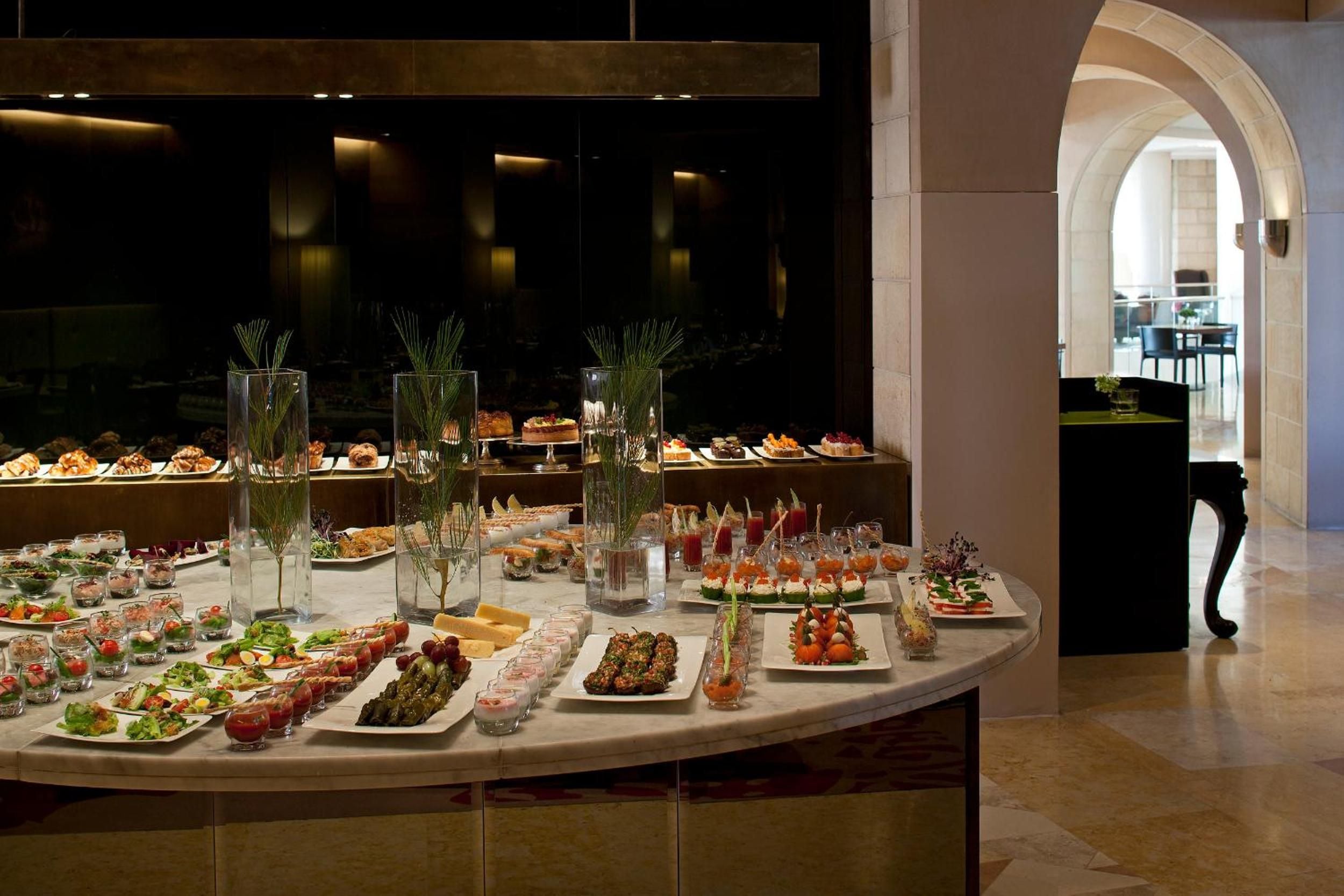 Artfully arranged appetizers and desserts create an elegant curved display, where tall grass arrangements and warm lighting transform dining into theater at this sophisticated Jerusalem retreat.