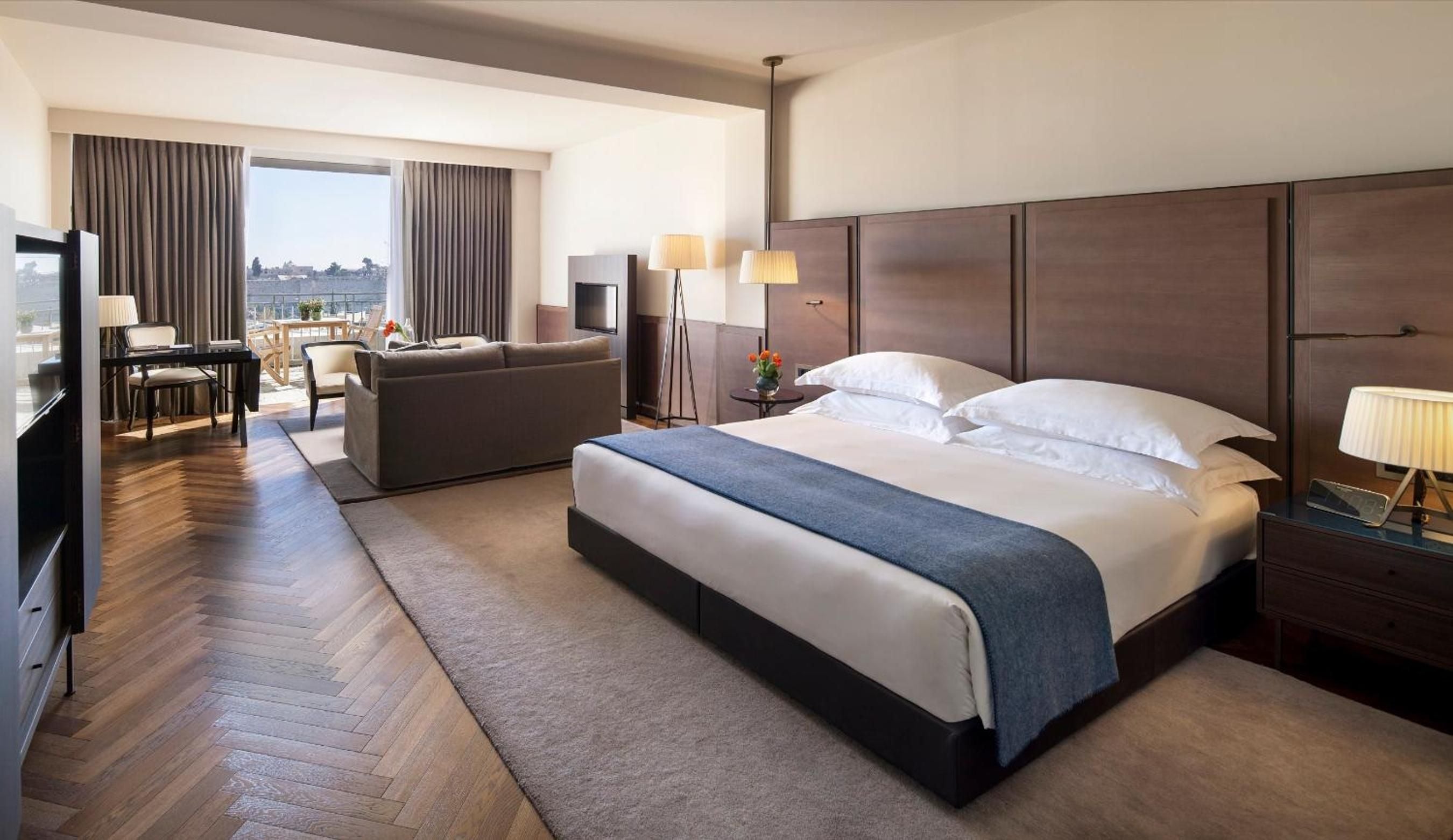 Warm wood floors and contemporary furnishings create an inviting sanctuary, where floor-to-ceiling windows frame Jerusalem's ancient skyline beyond flowing curtains.