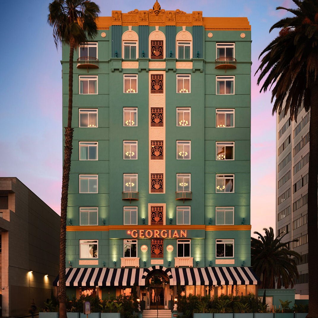Art Deco elegance meets California cool as the jade facade glows against twilight sky, striped awnings beckoning beneath swaying palms in Santa Monica.