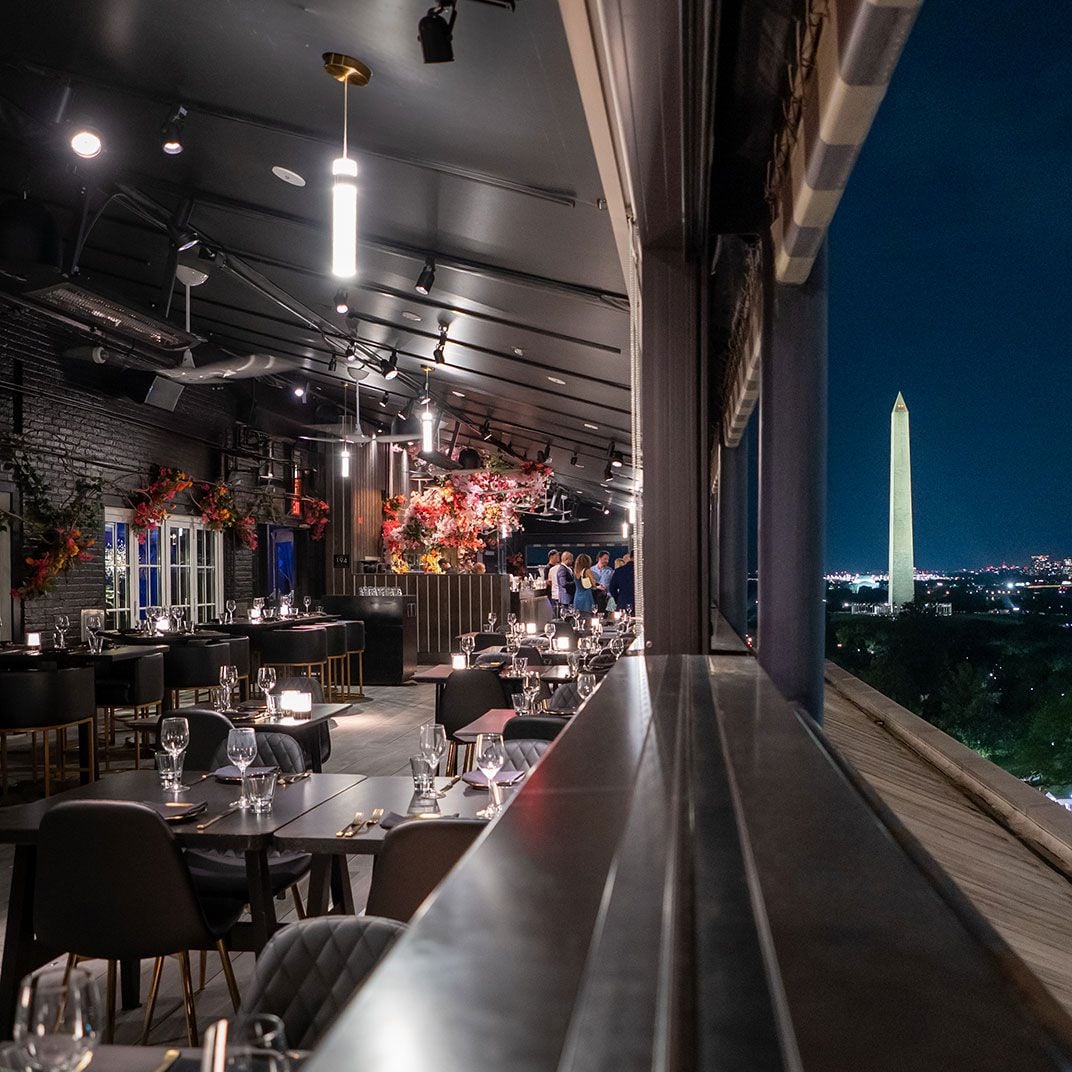 Floor-to-ceiling windows frame the illuminated Washington Monument as diners savor refined cuisine amid exposed brick walls and autumn florals at this elevated sanctuary.
