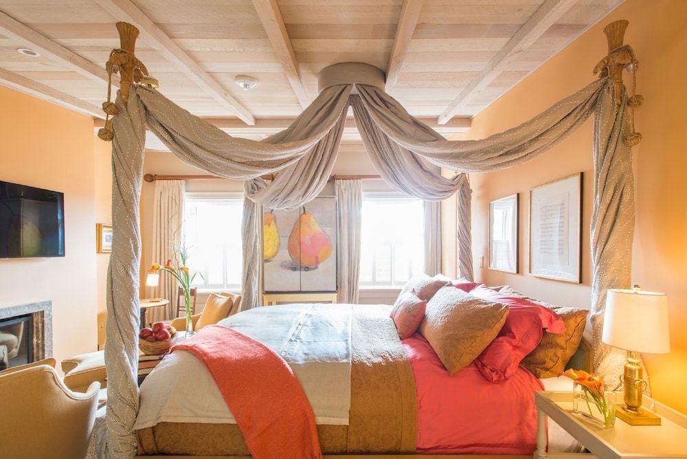 Draped canopy posts frame a dreamy retreat where coral-hued linens complement exposed beam ceilings, while golden hour light streams through oversized windows overlooking the city.