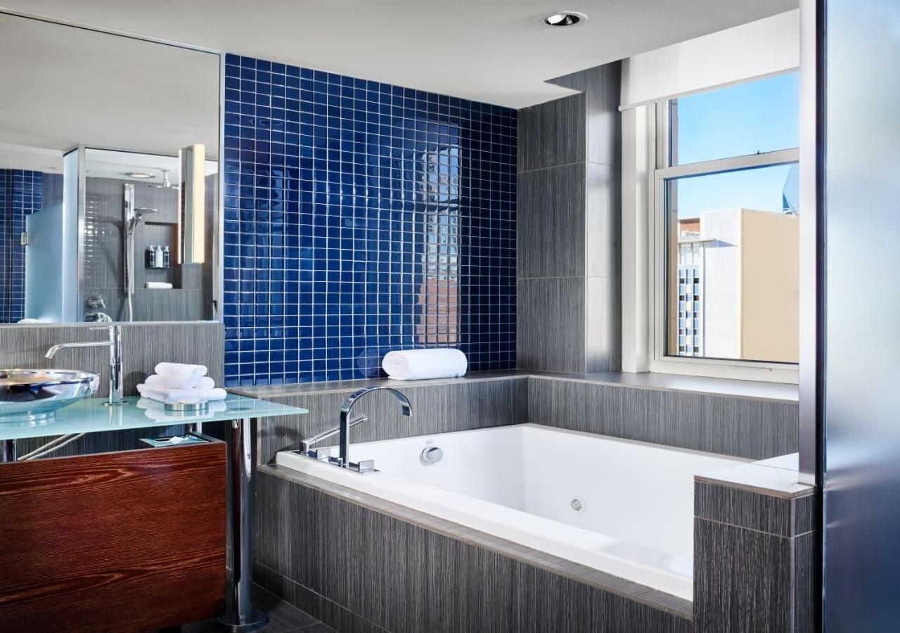 Cobalt mosaic tiles create a striking backdrop in this corner suite bathroom, where soaking tub meets city skyline through floor-to-ceiling windows.