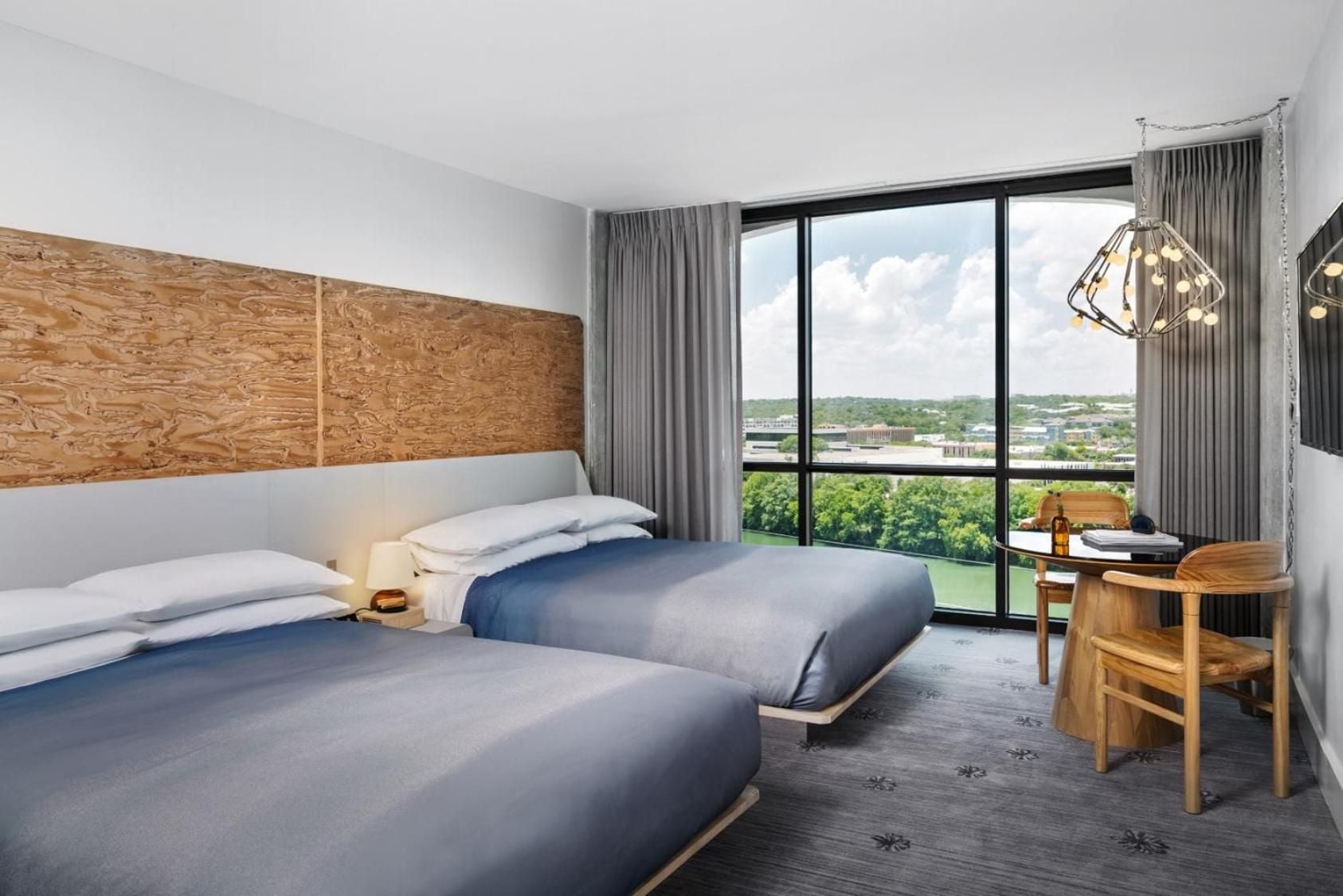 Warm wood headboards and industrial pendant lights create modern comfort in double-bed rooms, where floor-to-ceiling windows frame Austin's green canopy stretching toward downtown's skyline.