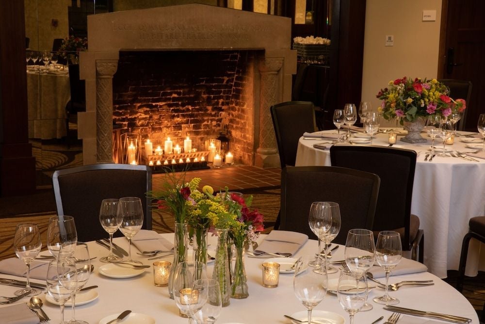 Candlelit tables arranged before a stone fireplace create an intimate dining atmosphere, where flickering flames dance against exposed brick and fresh florals add seasonal color.