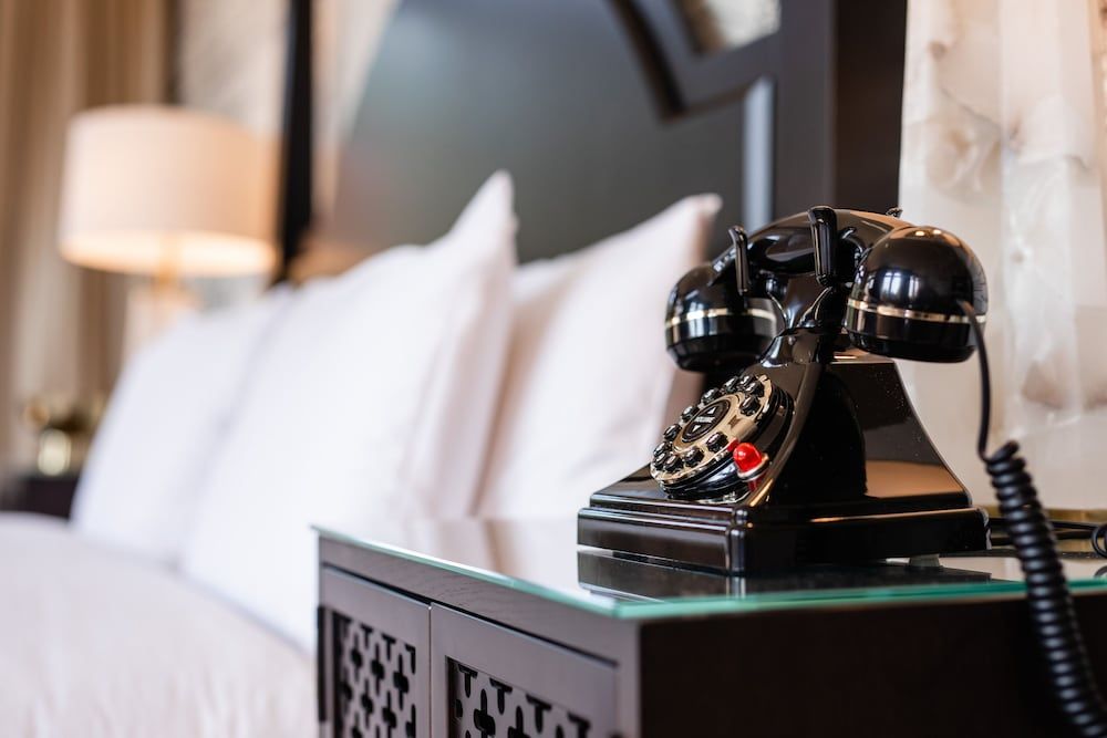 A vintage rotary telephone adds nostalgic charm to elegantly appointed guest rooms, where classic black lacquer meets crisp white linens and warm ambient lighting.