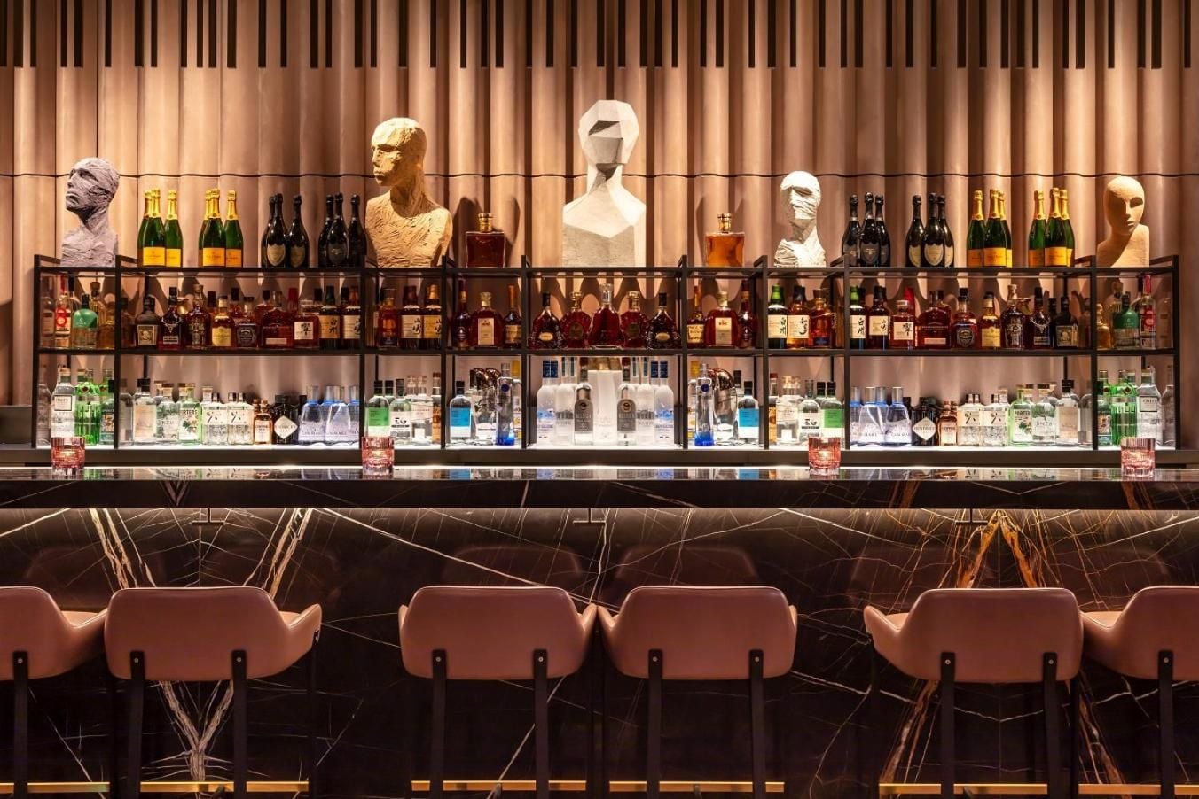 Sculptural busts and premium spirits create an art gallery atmosphere behind the marble bar, where copper-toned chairs invite intimate conversations over expertly crafted cocktails.