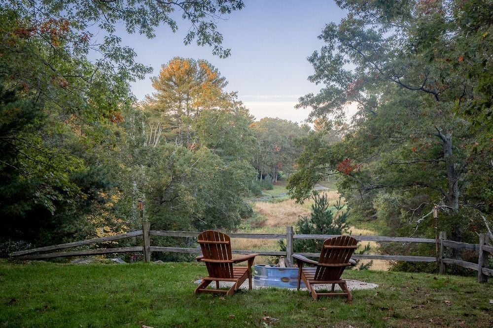 Two Adirondack chairs invite quiet contemplation overlooking autumn meadows, where split-rail fencing frames rolling hills dotted with maples turning golden in the fading light.