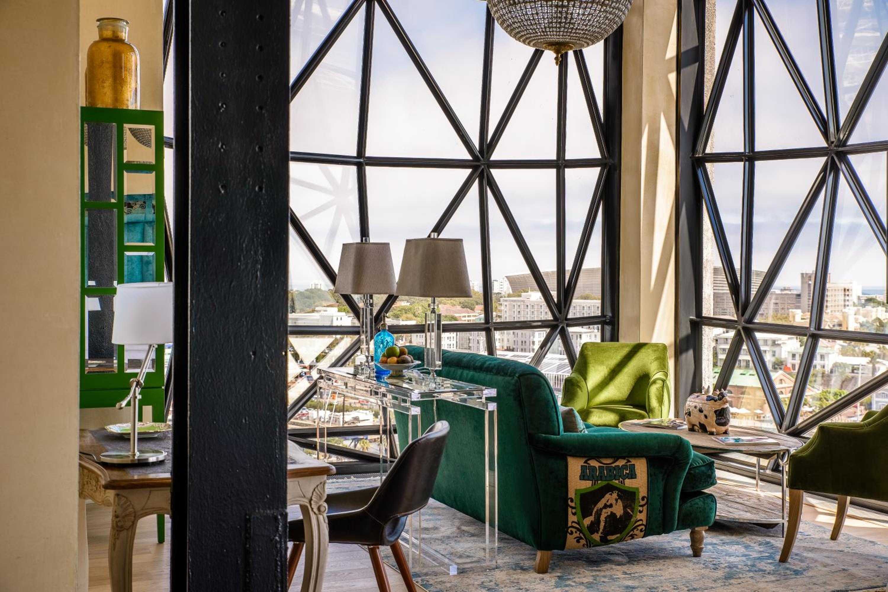 Emerald velvet armchairs and geometric windows frame Cape Town's cityscape in this striking suite, where industrial beams meet luxurious comfort beneath a glittering chandelier.