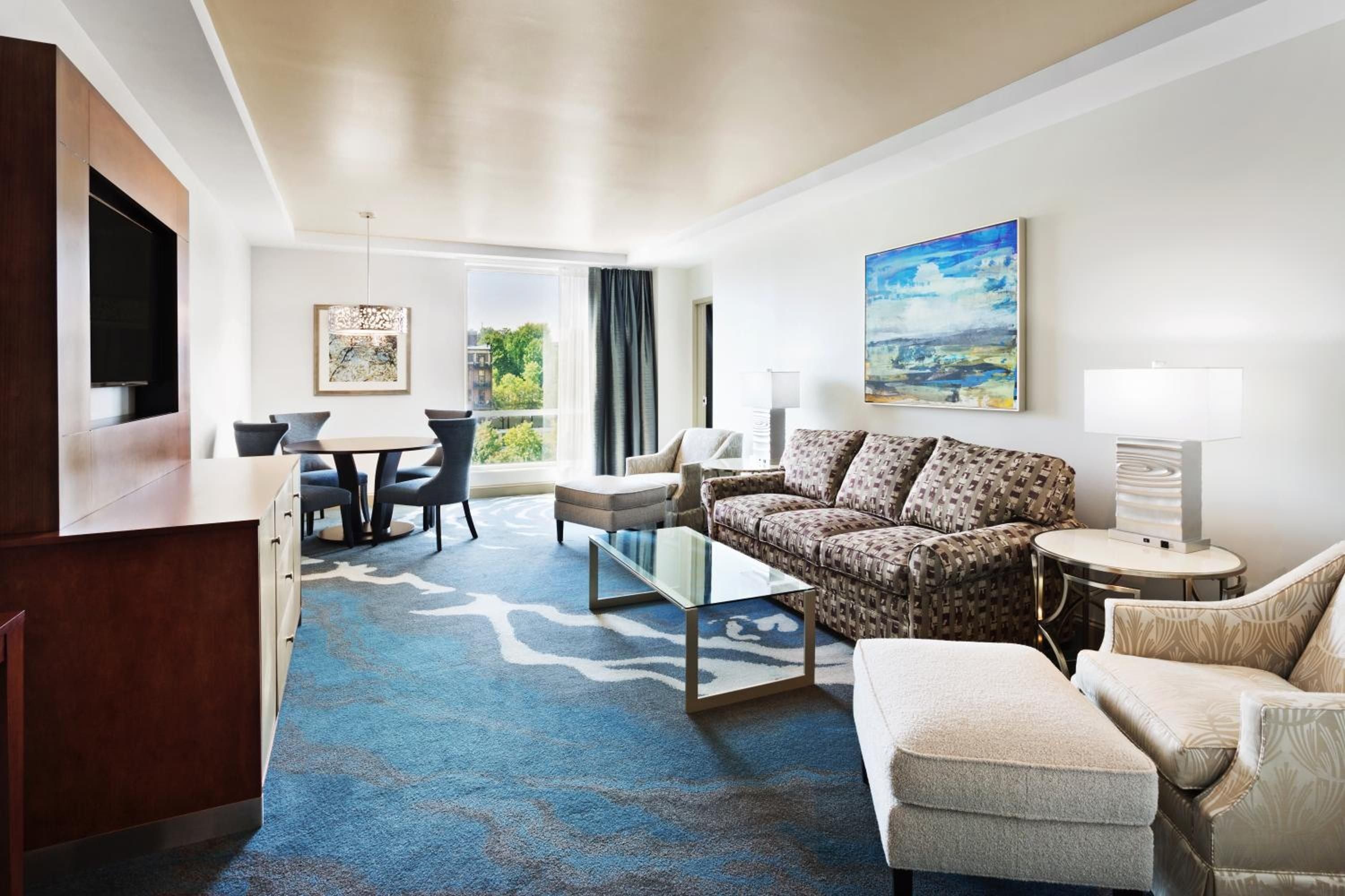 Geometric patterns dance across plush blue carpeting while natural light streams through floor-to-ceiling windows, creating an inviting residential-style living space with thoughtful artistic touches.