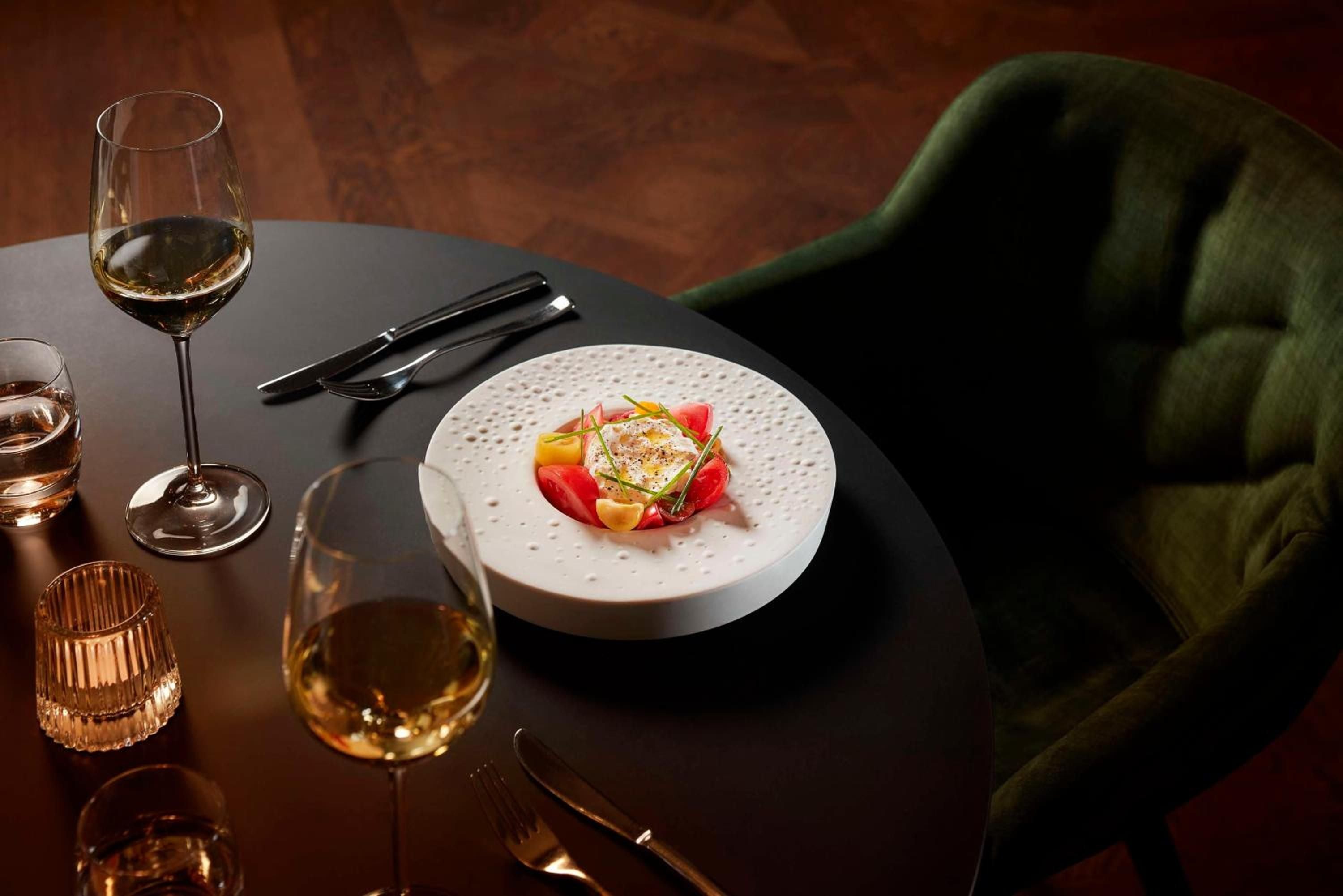Artfully plated tomato and cheese creation sits alongside amber wine on dark marble, illuminated by warm candlelight in the hotel's intimate dining space.