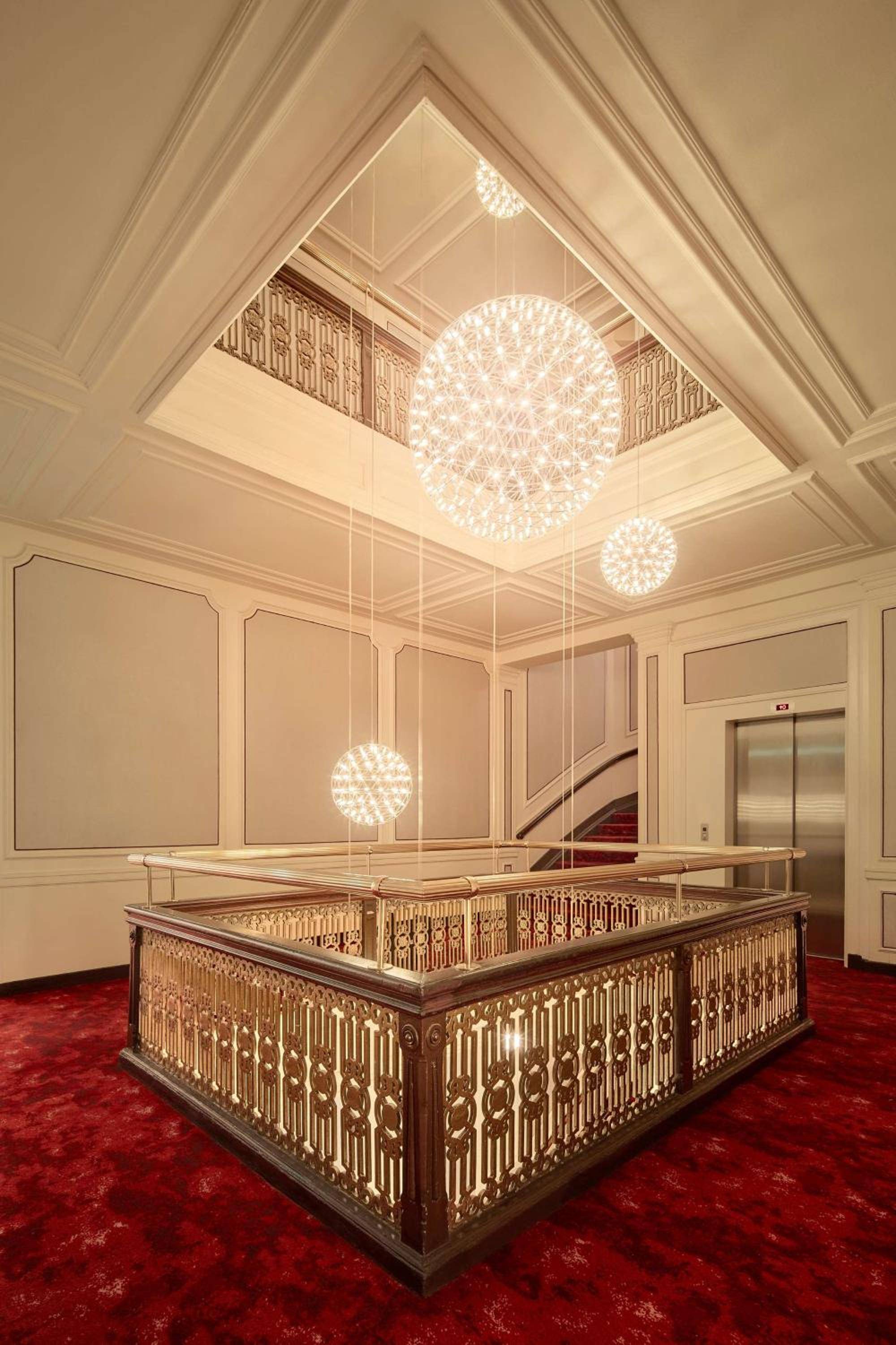Ornate golden balustrades frame a dramatic light well where crystalline orb chandeliers float like celestial bodies above plush crimson carpeting.