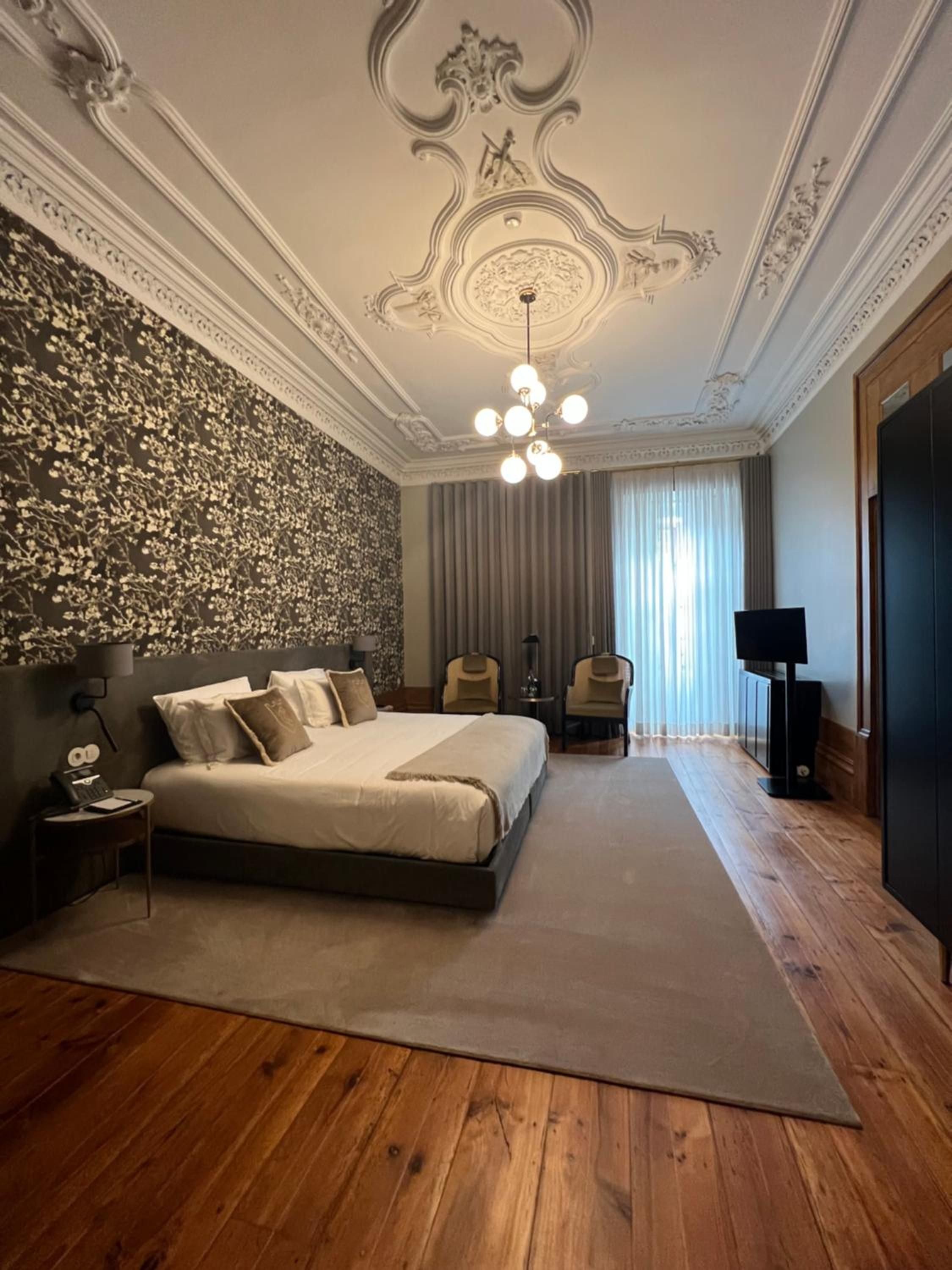 Ornate ceiling moldings and contemporary furnishings create an elegant contrast in this sophisticated guest room, where golden wallpaper catches warm light from the vintage chandelier.