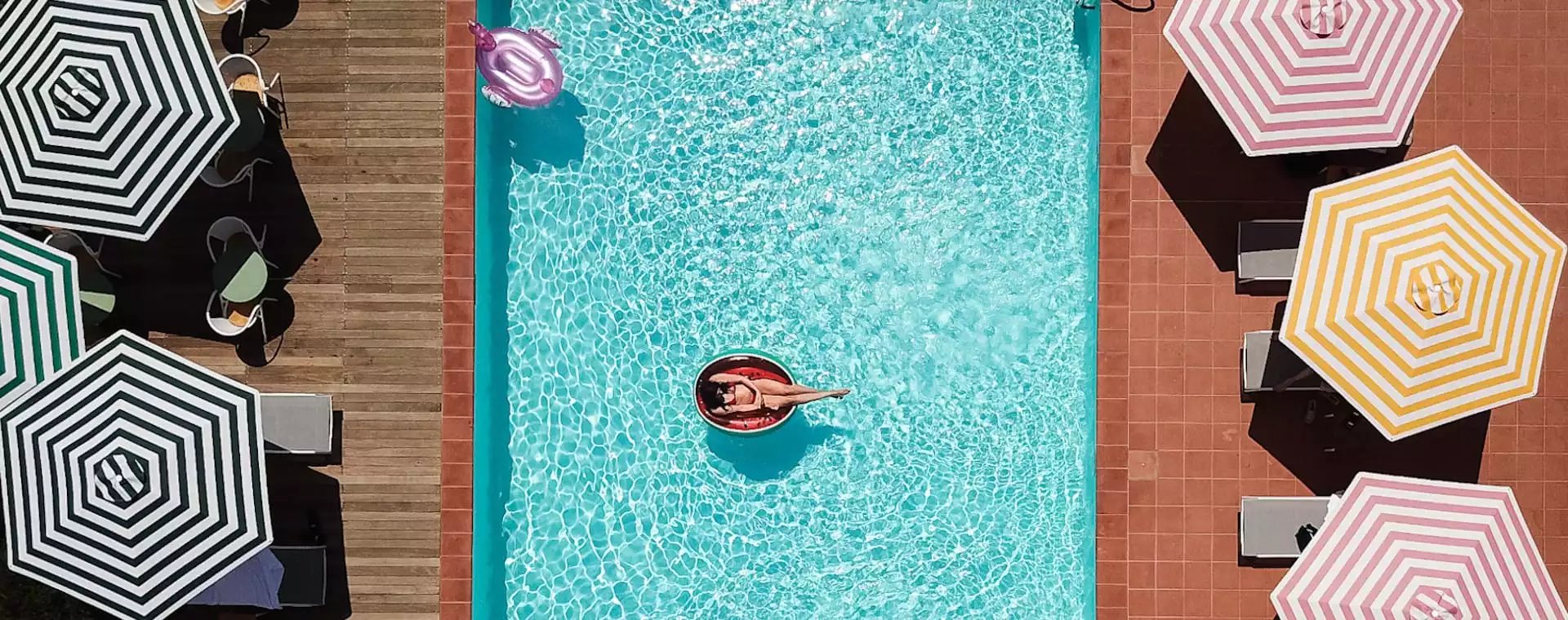 Striped umbrellas cast geometric shadows around Villa Lena's turquoise pool, where guests float lazily on watermelon-red inflatables beneath the Tuscan sun.