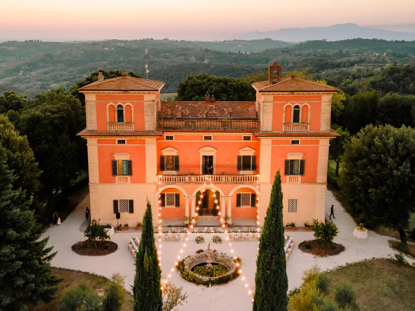 Villa Lena's coral-hued facade glows against Tuscan hills at golden hour, its symmetrical towers and cypress-lined courtyard fountain creating an enchanting Italian countryside tableau.