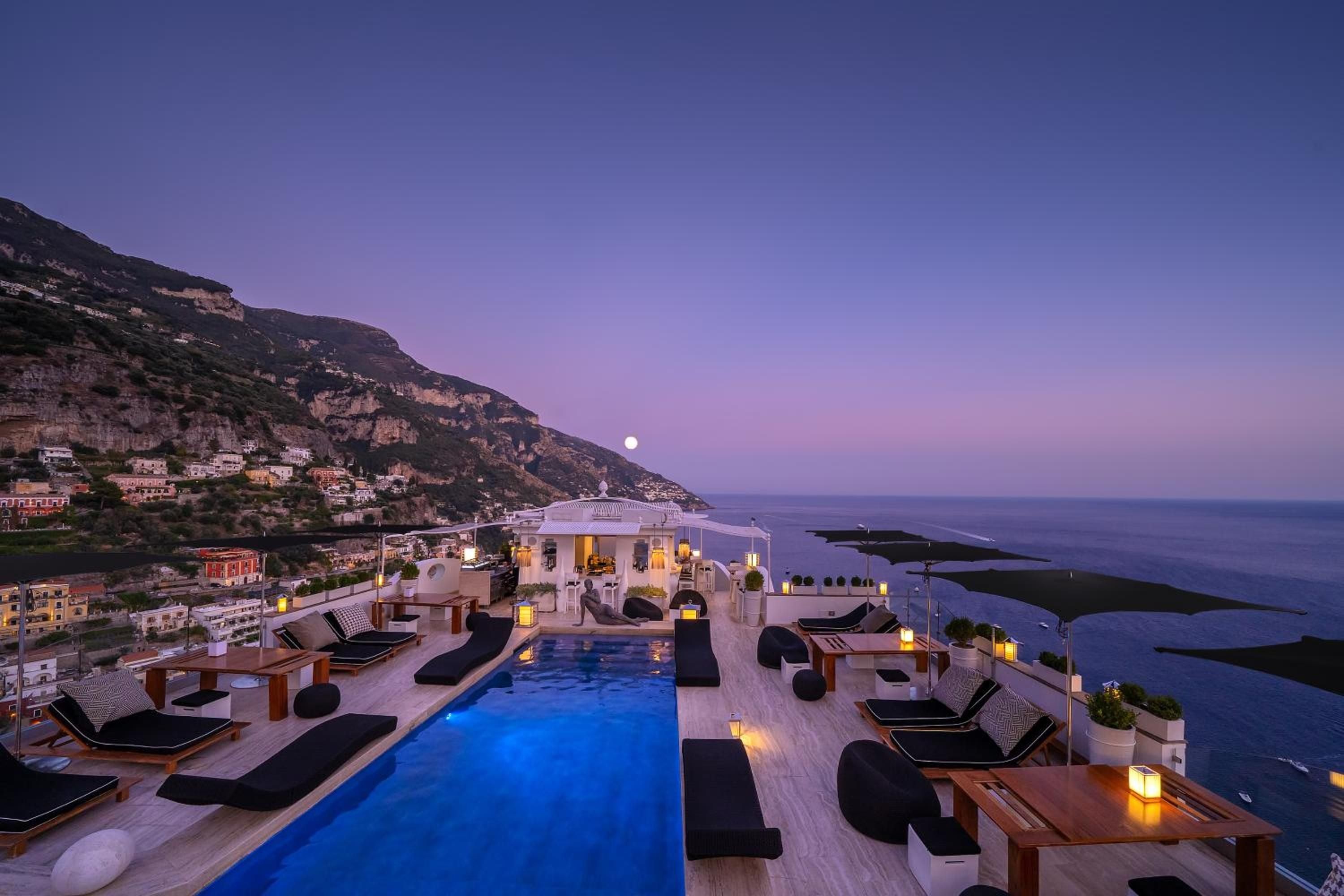 Infinity pool glows sapphire against twilight skies as lanterns flicker across the terrace, framing Positano's dramatic coastline and the rising moon beyond.
