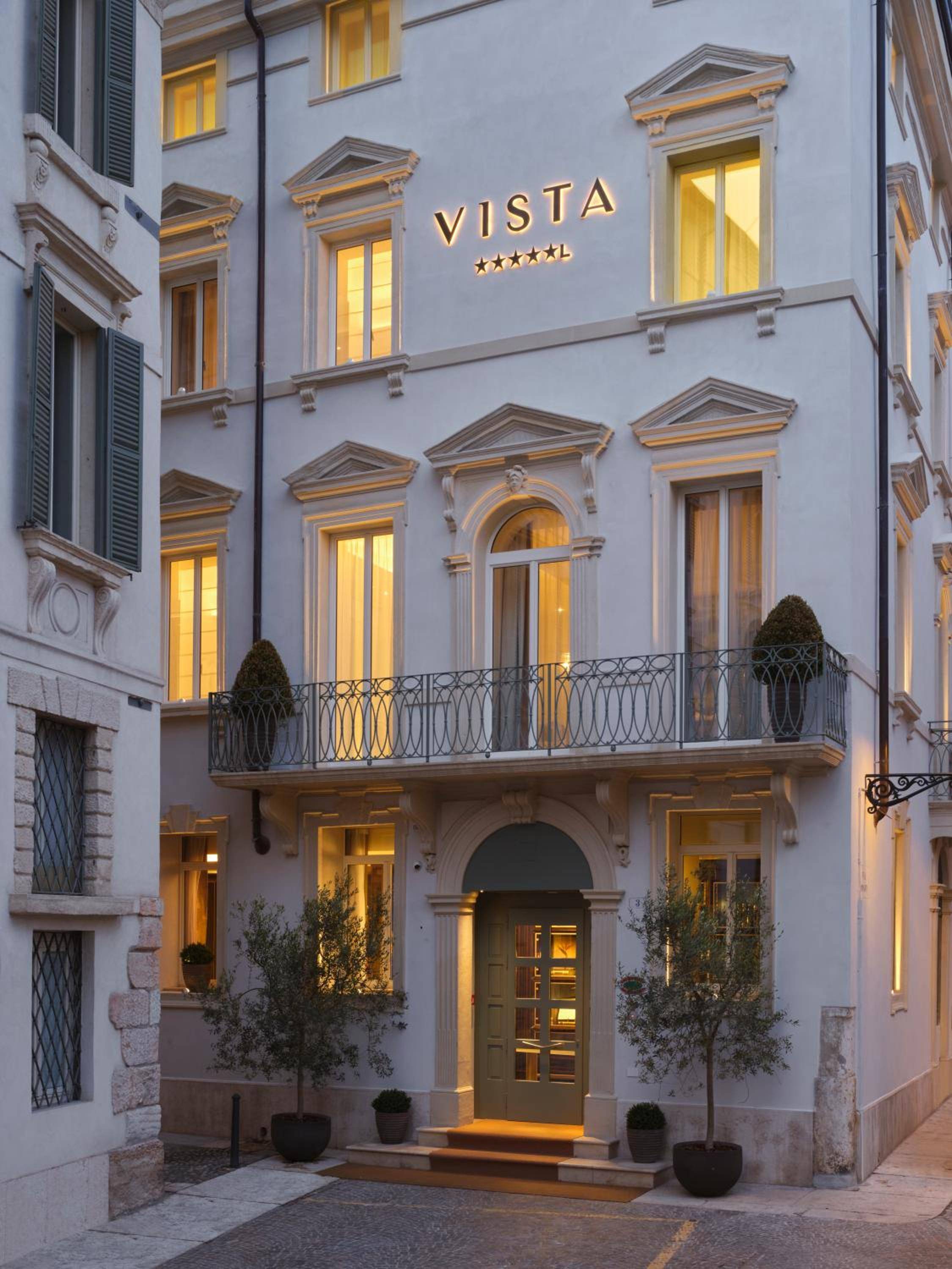 Warm golden light spills from Vista Verona's classical windows at dusk, where wrought-iron balconies and stone details create an intimate palazzo atmosphere on cobbled streets.