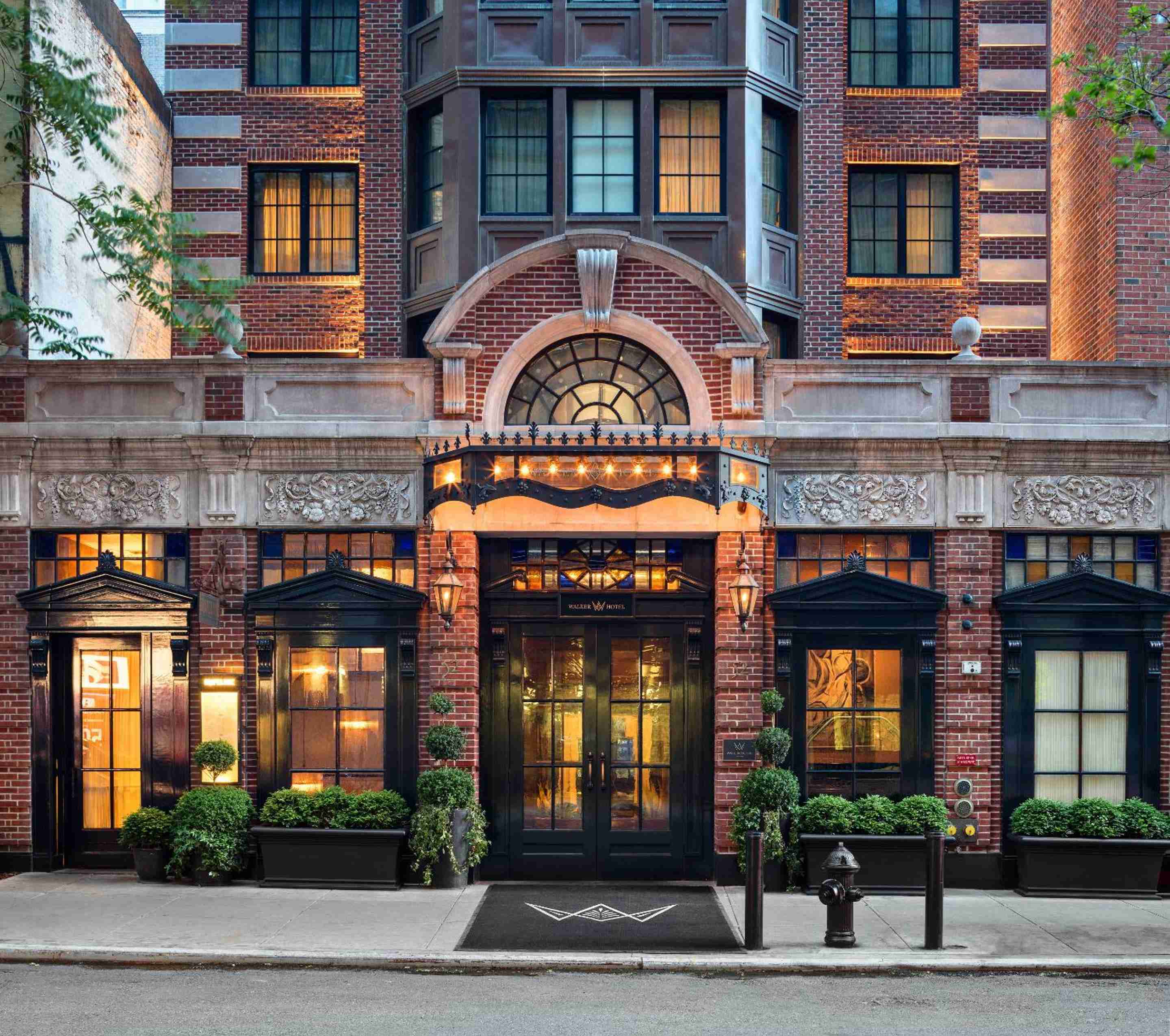 Warm amber light spills from arched windows above the elegant entrance, where ornate stonework and manicured topiaries create an inviting Greenwich Village sanctuary.