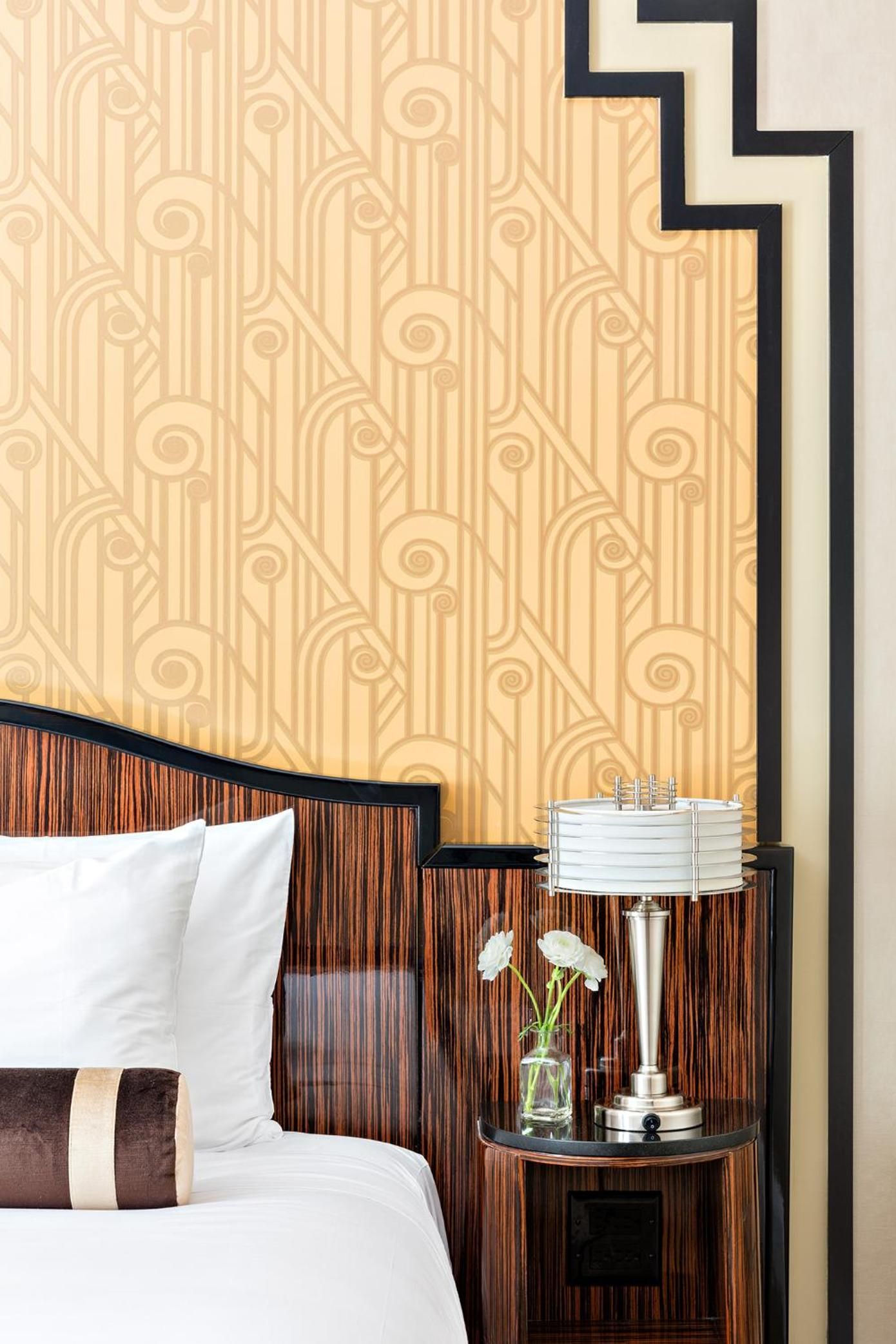 Art Deco wallpaper in warm gold tones creates an intimate backdrop for rich zebra wood furnishings and crisp white linens in this thoughtfully appointed guest room.