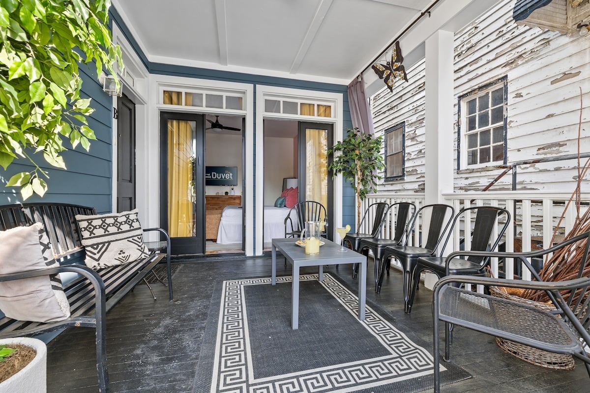 CAPTION: Nestled in Charleston, "Property" exudes Southern charm with its colonial architectural style, featuring a serene porch adorned with classical wood and wicker seating. The rich blue and white facade offers a striking contrast against the rustic weathered textures of adjacent buildings. Lush greenery and modern decor elements create a tranquil and inviting atmosphere. Ideal for travelers seeking relaxation, this luxurious setting is perfect for unwinding and enjoying Charleston's historic beauty.