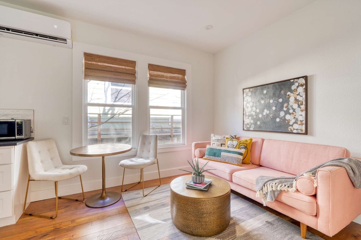 CAPTION: 
Property in Portland showcases a chic, contemporary design with a warm and inviting atmosphere. The room features a soft color palette with pastel pinks, creamy whites, and natural wood tones. Key visual elements include a plush pink sofa, abstract wall art, and stylish metallic accents. Large windows flood the space with natural light, creating a serene environment perfect for luxury travelers seeking a modern yet cozy retreat.