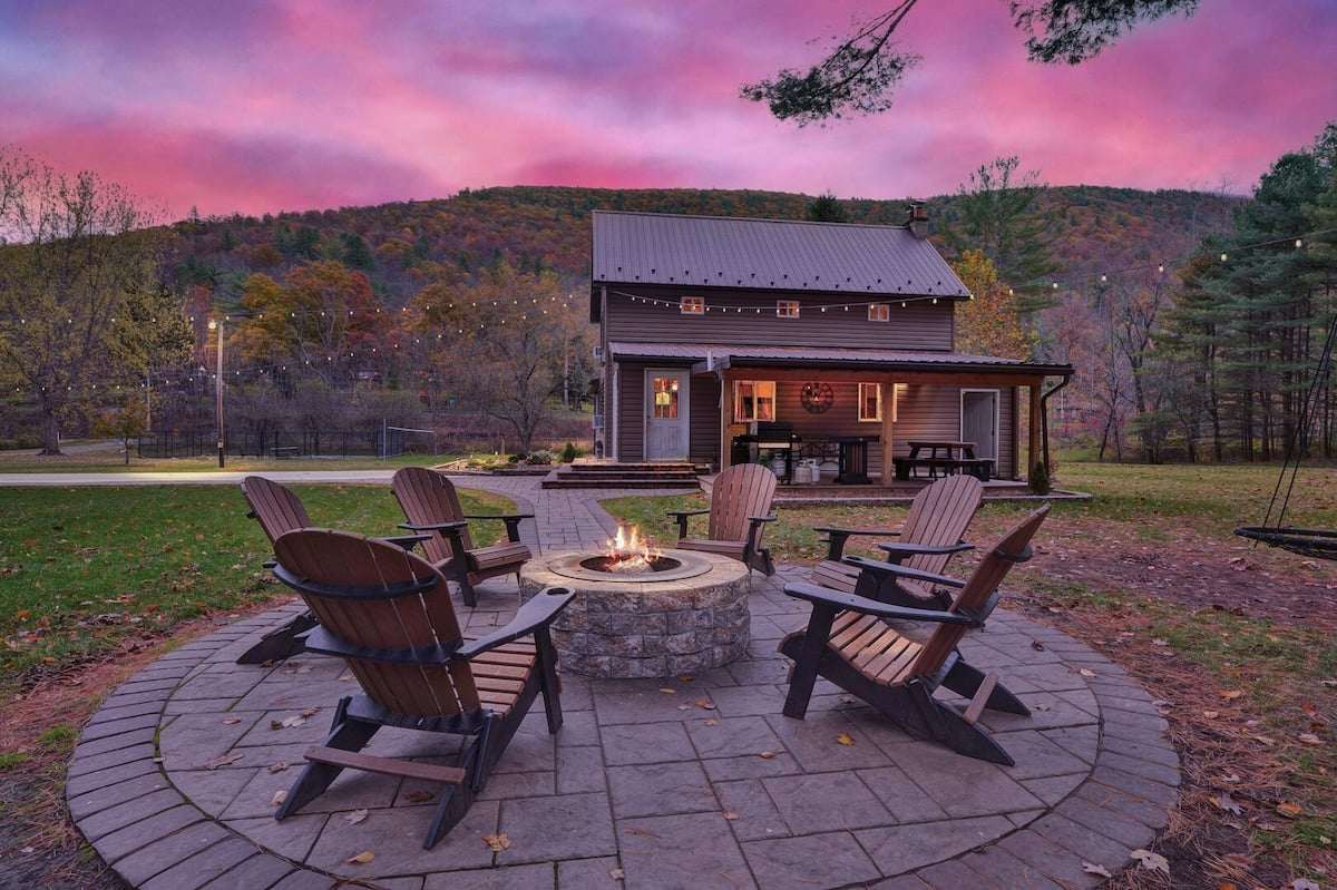 **CAPTION:** The "Property" in Cedar Run showcases rustic charm with its cozy, cabin-style architecture surrounded by lush woodland. This inviting retreat features a central stone fire pit, perfect for evening gatherings under the enchanting string lights. The tranquil atmosphere is enhanced by a vivid sunset palette of pinks and purples, offering a serene escape. Ideal for travelers seeking a peaceful, nature-centric getaway, this property promises a memorable stay with modern comforts.