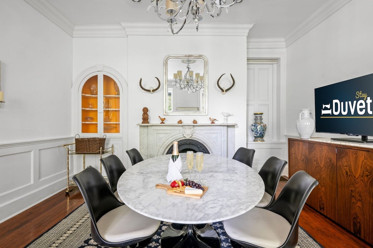 CAPTION: Nestled in Charleston, "Property" exudes a sophisticated mid-century modern charm, featuring a luxurious dining area with a sleek marble table. The room's elegant design is highlighted by a soft monochrome color palette, accented by rich wooden floors and a chic chandelier. With its blend of contemporary style and timeless elegance, this inviting space offers a serene atmosphere perfect for discerning travelers seeking a refined retreat.