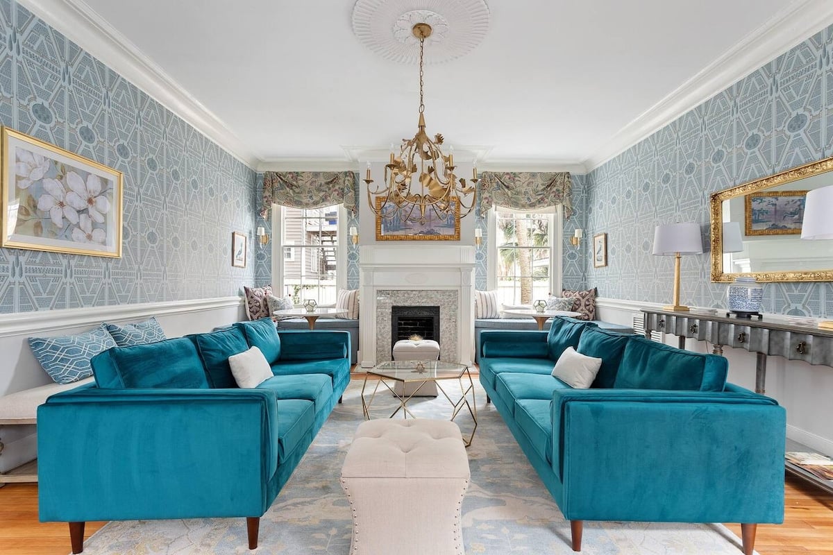 CAPTION: Nestled in the heart of Charleston, "Property" exudes Southern elegance with its sophisticated Georgian architectural style. The interior features a serene atmosphere, highlighted by its luxurious teal velvet sofas and an ornate gold chandelier. The room's blue and white color palette, complemented by floral accents and intricate wallpaper, creates a harmonious and inviting ambiance. Ideal for discerning travelers seeking a blend of classic charm and modern comfort, this opulent residence captures the essence of refined luxury.