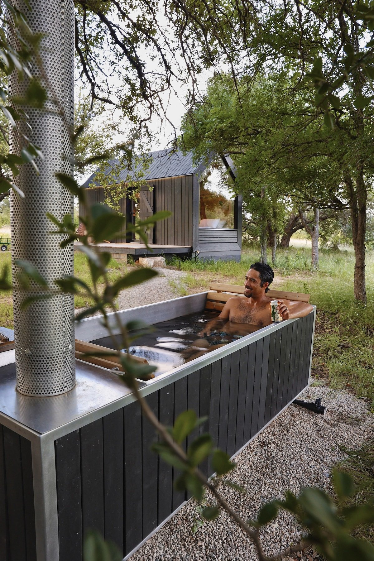 CAPTION: Nestled in the serene landscape of Round Mountain, "Property" exudes rustic elegance with its modern cabin architecture. The inviting hot tub set against a backdrop of lush greenery creates a tranquil retreat perfect for relaxation. Its sleek black and wood design harmonizes with the natural surroundings, offering a calming escape for travelers seeking an intimate connection with nature. The warm, earthy tones and peaceful atmosphere make it a desirable destination for relaxation and rejuvenation.