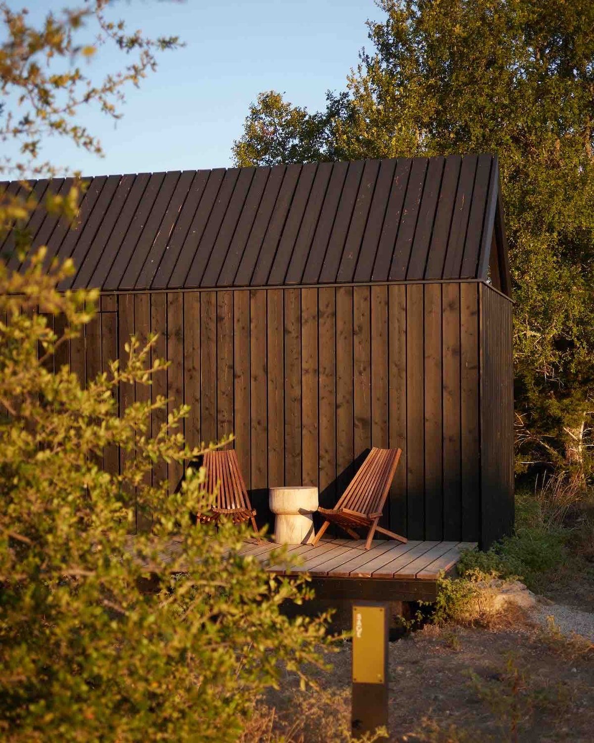 CAPTION:  
"Property in Round Mountain showcases a modern rustic architectural style, seamlessly blending into its natural surroundings. The dark wooden exterior exudes a warm, welcoming atmosphere, inviting travelers to relax. Two elegant wooden chairs and a unique stone table on the deck offer a perfect spot for unwinding amidst nature. The earthy color palette of dark browns and greens enhances the serene mood, appealing to those seeking a tranquil retreat."