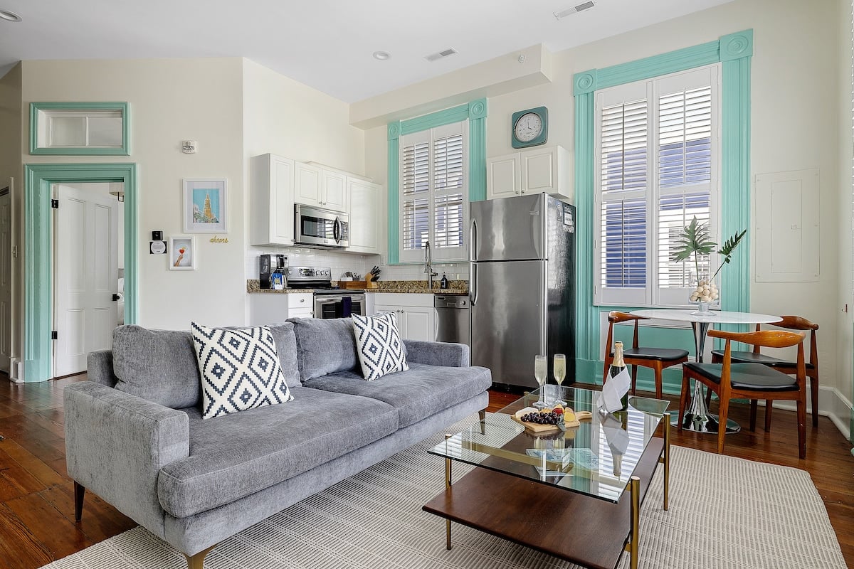 CAPTION: Nestled in the heart of Charleston, "Property" exudes a modern urban charm with its contemporary interior design. The open-plan living space features sleek silver appliances and plush seating, accented by a crisp white and soft teal color palette. Wooden floors add warmth, while ample natural light enhances the airy atmosphere. Perfect for discerning travelers seeking comfort and style, this luxury retreat offers a serene escape amidst the bustling city.