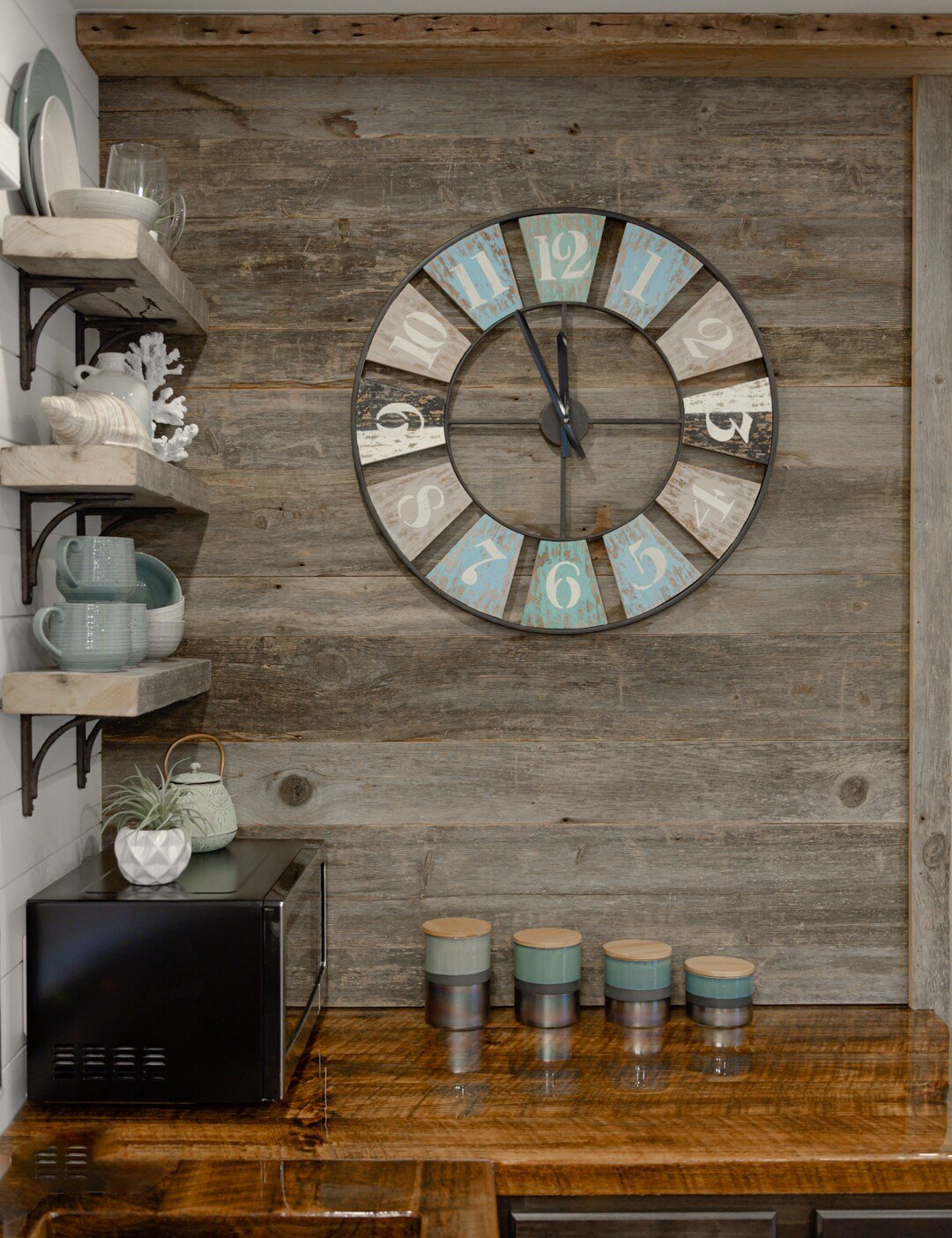CAPTION: 
The "Property" in Waco exudes rustic charm with its farmhouse-inspired interior. The focal point is a vintage-style clock against a backdrop of weathered wood paneling, evoking a cozy, nostalgic atmosphere. The earthy color palette of browns, creams, and teal blue enhances its inviting warmth. Open shelves with curated ceramics and sleek wooden countertops add modern sophistication, appealing to travelers seeking a stylish yet homey retreat.
