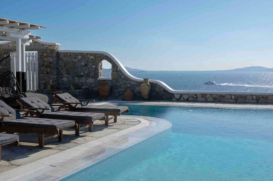 **CAPTION**: Nestled in the serene Agios Andreas Korakochoriou, GR, "Property" embodies Mediterranean elegance with its classic stone architecture and breathtaking sea views. The tranquil atmosphere is enhanced by the infinity pool blending seamlessly with the horizon, offering a luxurious escape. The warm hues of the sunlit stone, contrasted with azure waters, create a stunning visual palette. This exquisite retreat promises ultimate relaxation and an unparalleled experience for discerning travelers.