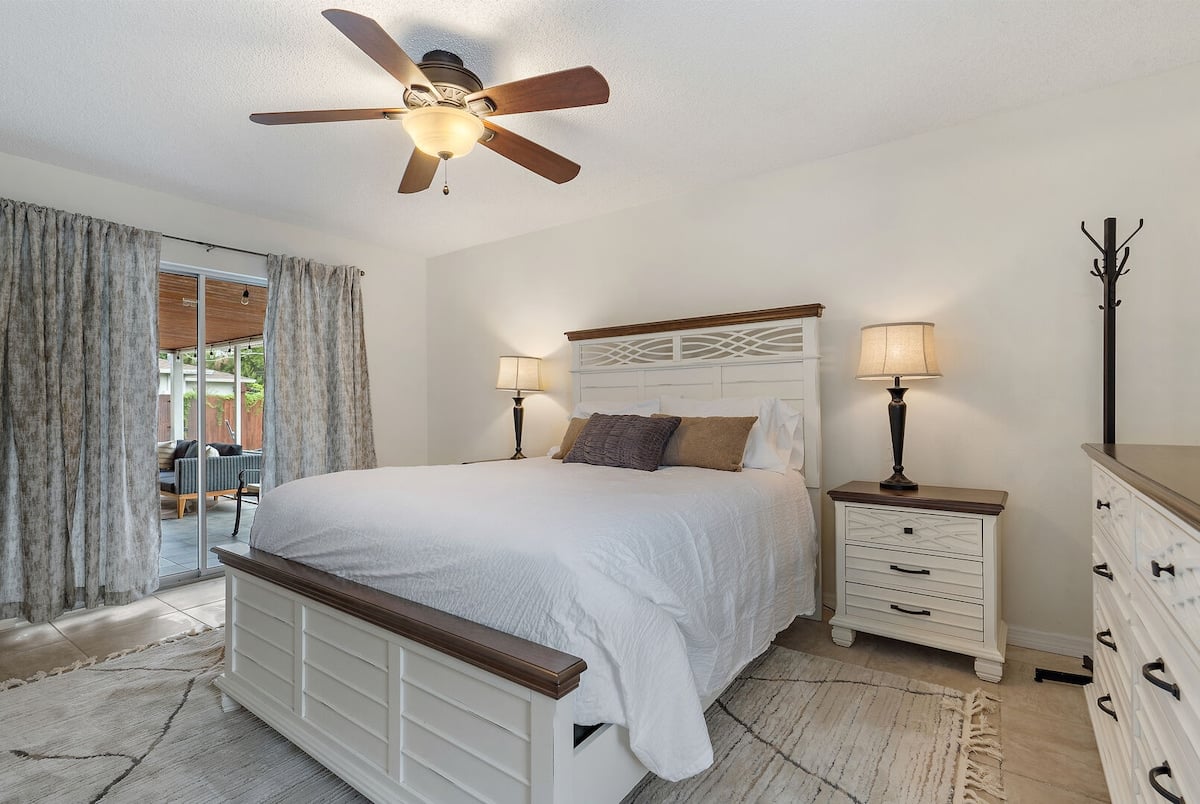 **CAPTION**: Nestled in Sarasota, "Property" exudes a warm, coastal charm with its modern yet rustic architectural style. The inviting bedroom features a neutral color palette with soft whites and earthy accents. A wooden ceiling fan and textured, light-filtering curtains add a touch of comfort and elegance. Perfect for discerning travelers seeking a relaxed yet refined atmosphere, this serene space offers a tranquil retreat, seamlessly blending indoor and outdoor living with views of the lush surroundings.
