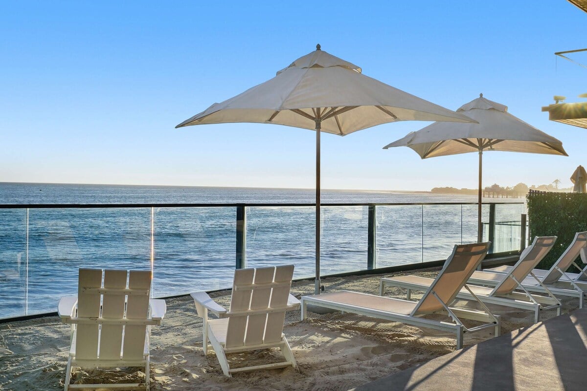 CAPTION: 
Property in Malibu epitomizes coastal luxury with its modern, open-air design and breathtaking oceanfront views. Located directly on the sandy shores, the property features sleek glass railings and chic white lounge chairs, all against a serene blue ocean backdrop. Ideal for sun-seekers and relaxation enthusiasts, its minimalist aesthetic and calming color palette invite travelers seeking an exclusive beachfront retreat to unwind and enjoy the tranquility of the Pacific coastline.