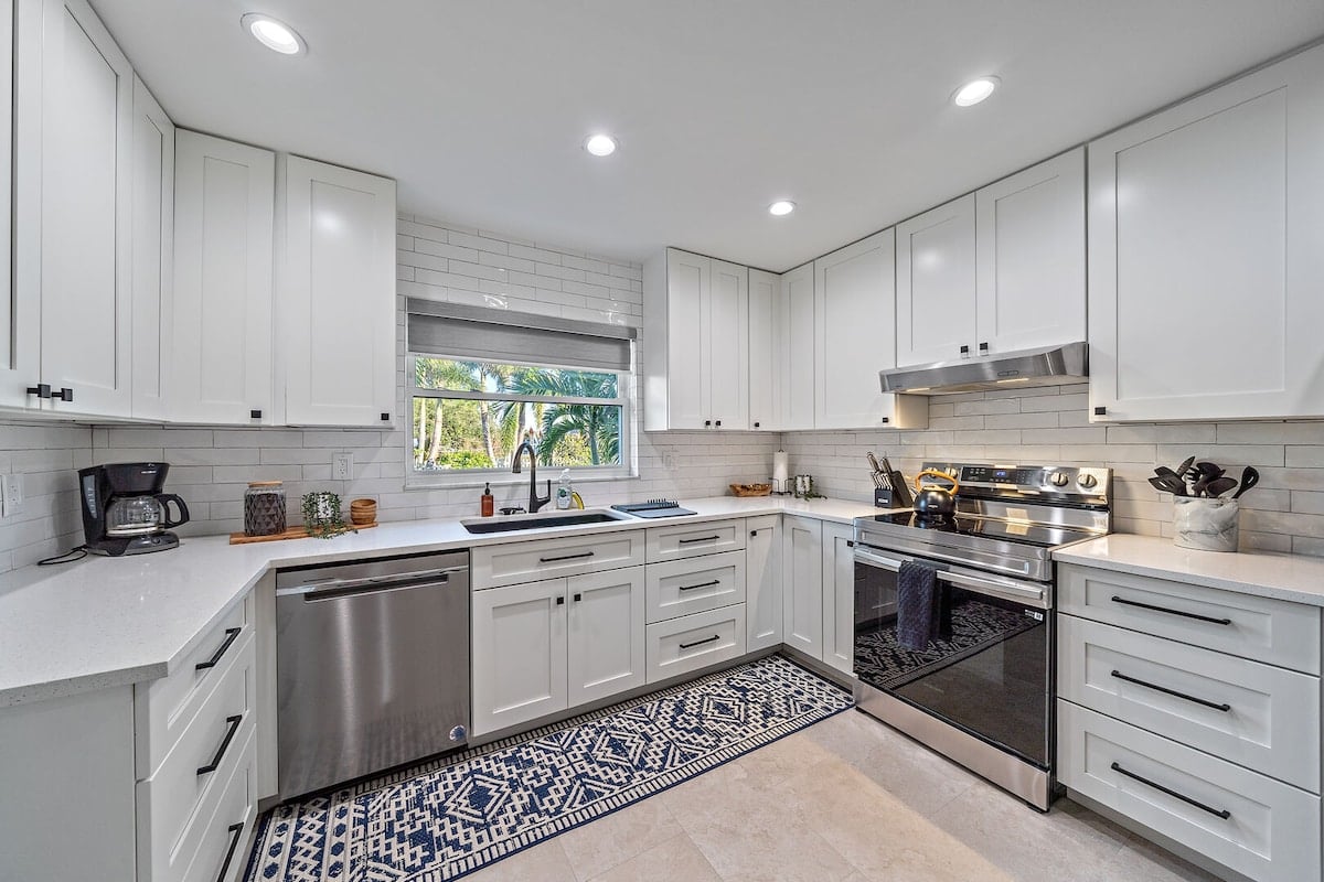 CAPTION: Nestled in Sarasota, "Property" exudes modern elegance with its sleek, contemporary kitchen design. The space features pristine white cabinetry paired with a cool gray countertop, complemented by stainless steel appliances that add a touch of sophistication. Sunlight filters through a large window, highlighting the tranquil palm views outside. A patterned blue and white rug adds warmth and charm, making it a perfect setting for travelers seeking luxury and style.