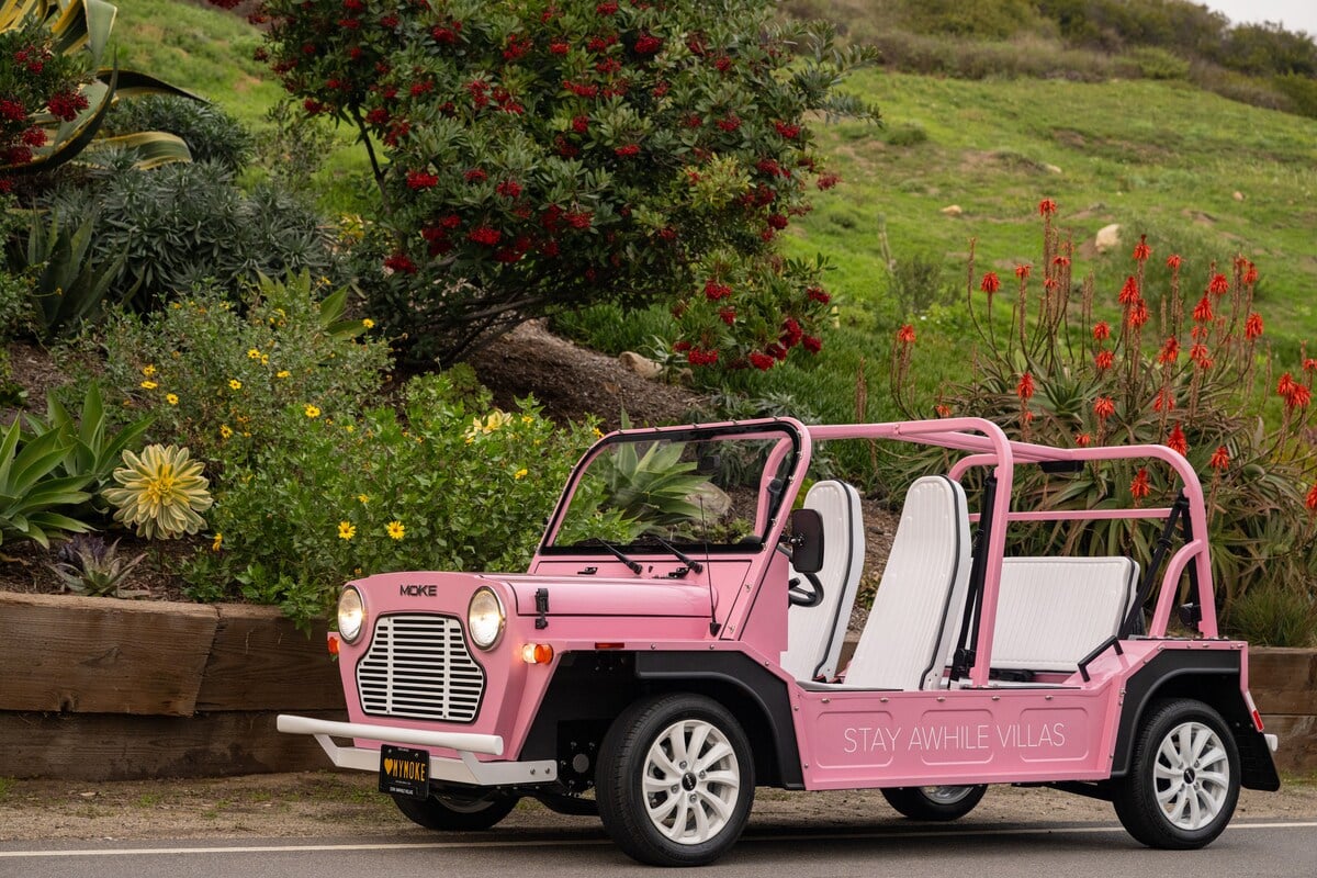 **CAPTION**: Discover "Property," a luxurious haven in Malibu that blends modern comfort with vibrant coastal charm. This pink open-air mini-moke exudes a playful, adventurous atmosphere, ideal for exploring the scenic seaside surroundings. The lush greenery and blooming flora enhance the vivid color palette, creating an inviting retreat for travelers seeking relaxation and adventure. The sleek, minimalist design of the vehicle complements the exquisite backdrop, making "Property" a standout destination for luxury seekers.