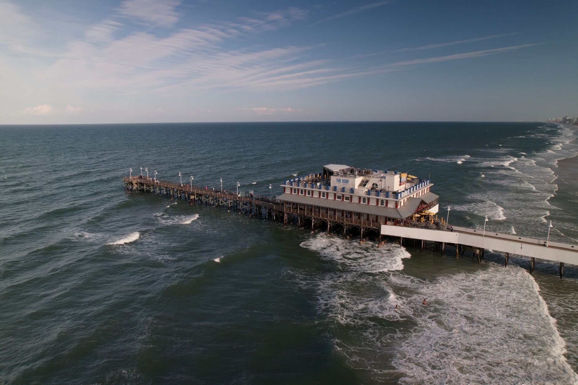 Aerial view of a pier extending into the ocean with waves crashing gently along the shore.