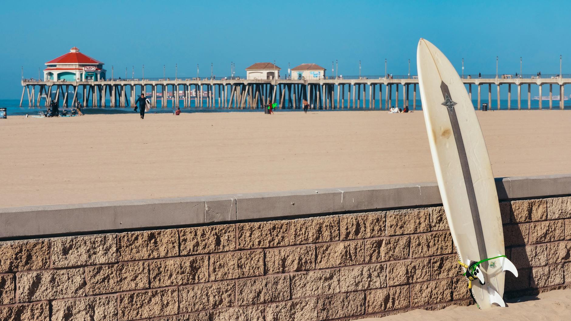 Sunny day at Huntington Beach with a surfboard resting on a brick wall and iconic pier in the background.
