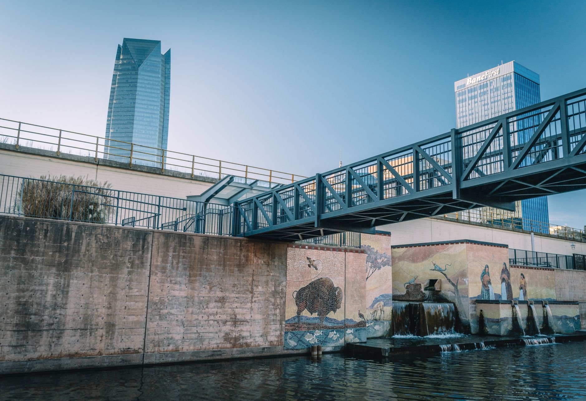View of Oklahoma City's canal featuring a footbridge, murals, and skyscrapers in the background.